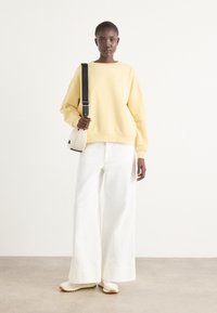 Gul oversized sweatshirt med rund halsringning, parat med vida vita byxor. Modellen bär en liten krämfärgad crossbody-väska och har på sig ljusa skor.