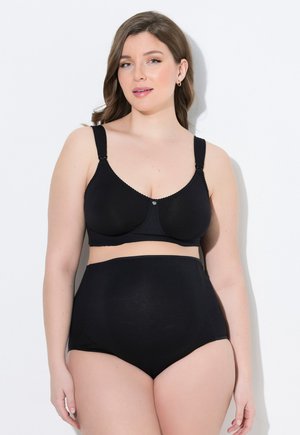 Femme portant un soutien-gorge de maternité noir et une culotte de maternité noire taille haute, debout devant un fond blanc.