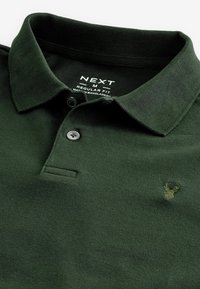 Polo vert foncé avec deux boutons noirs, col pointu, petit logo cerf brodé et étiquette imprimée indiquant la taille et la coupe à l'intérieur du col.