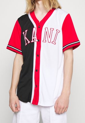 Personne portant un maillot de baseball rouge, noir et blanc à boutons avec les lettres "KANI" sur le devant, associé à un pantalon blanc.