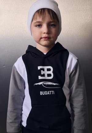 Bambino piccolo con berretto bianco e felpa nera e grigia con cappuccio con logo Bugatti e silhouette di un'auto, in piedi su uno sfondo sfumato neutro.