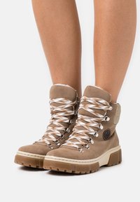 Des bottes de randonnée en suède marron présentent un col rembourré, des lacets blancs et marron, des œillets en métal et une semelle en caoutchouc robuste avec une surface texturée offrant une bonne adhérence.