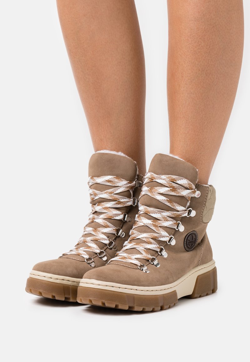 Des bottes de randonnée en suède marron présentent un col rembourré, des lacets blancs et marron, des œillets en métal et une semelle en caoutchouc robuste avec une surface texturée offrant une bonne adhérence.