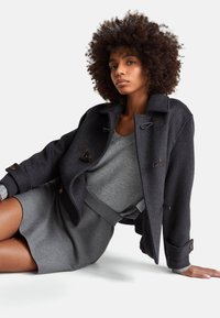 Dunkler Wollmisch-Peacoat mit Knebelverschlüssen, über einem hellgrauen Pullover getragen, kombiniert mit einem passenden grauen Rock. Insgesamt glatte Textur.