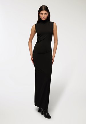 Abito maxi nero senza maniche con collo alto, caratterizzato da una silhouette aderente e una consistenza morbida, indossato con stivaletti neri con tacco.