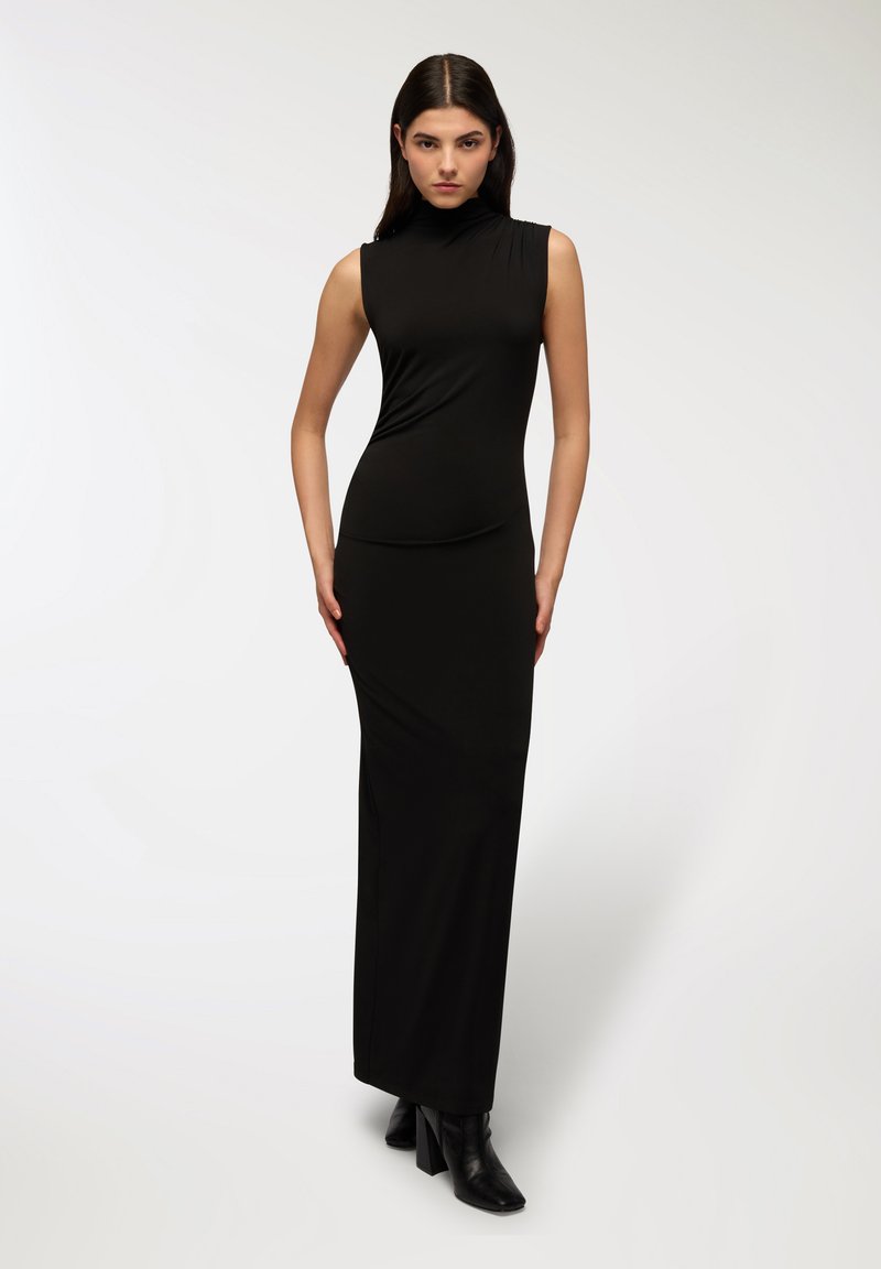 Abito maxi nero senza maniche con collo alto, caratterizzato da una silhouette aderente e una consistenza morbida, indossato con stivaletti neri con tacco.