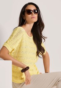 Geel gebreid sweater met korte mouwen, voorzien van opengewerkte patronen. Bruine zonnebril en een chunky bruine armband accentueren de look.