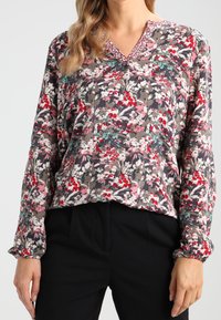 Mujer vestida con una blusa floral de mangas largas con flores rojas, rosas, blancas y turquesas, combinada con pantalones negros.