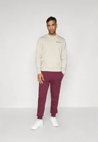 Sweat-shirt beige clair avec un petit logo sur la poitrine, associé à un pantalon de survêtement bordeaux fuselé. Des baskets blanches complètent la tenue.