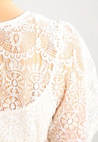 Vit spetsblus med intrikata blommönster och genomskinliga sektioner, med en strukturerad yta och rund halsringning.
