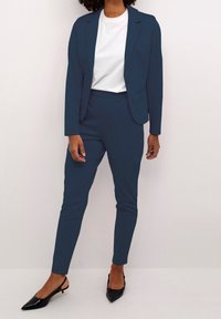 Granatowy garnitur ze slim-fit marynarką i spodniami, charakteryzujący się gładką teksturą. W zestawie z czarnymi czółenkami na szpilce i białym t-shirtem pod spodem.