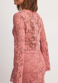 Pink lace dress featuring long sleeves, intricate floral patterns, and a fitted silhouette with a sheer back detail. Textured finish throughout.