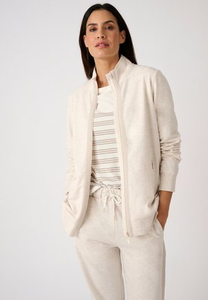 Donna con capelli scuri che indossa una giacca beige chiaro con zip sopra una camicia bianca a righe beige e pantaloni abbinati con coulisse, in piedi con le mani nelle tasche.