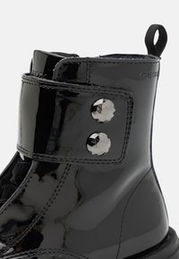 Stivale alla caviglia in pelle verniciata nera con una cintura in Velcro caratterizzata da due borchie decorative in argento, punta rotonda e suola texturizzata.