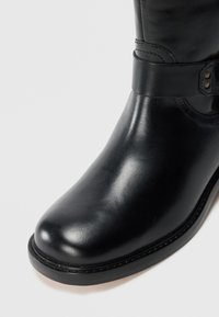 Botte en cuir noir à bout rond et finition lisse. Présente une sangle avec des détails en métal près de la cheville. Design minimaliste.