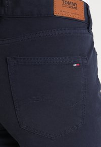 Jeans de mezclilla azul marino con una etiqueta de cuero marrón que dice "TOMMY JEANS", un bolsillo trasero con detalles de costura y acentos en rojo, blanco y azul.