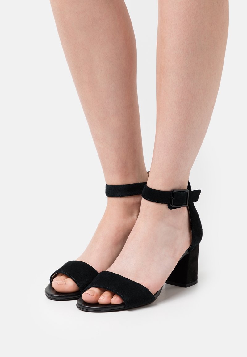 Jambes portant des sandales noires en daim à talons blocs ouvertes à l'avant avec des brides à la cheville et des boucles sur fond blanc.