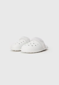 Vita slip-on klackar med runda ventilationshål och mjuk, fluffig vit foder runt öppningen, placerade sida vid sida på en enkel bakgrund.