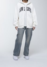 Sudadera blanca con cremallera y el texto "LOW LIGHT'S" en negro, combinada con vaqueros azules holgados y zapatillas blancas. Estilo casual y ajuste relajado.