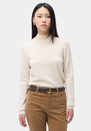 Junge Frau mit glatten dunklen Haaren, die einen cremefarbenen Rollkragenpullover, braune Hosen, einen dunklen Gürtel und goldene Creolen trägt, stehend vor einem einfarbigen Hintergrund.