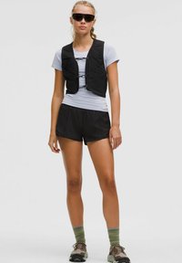 Vrouw in een zwart tactisch vest over een wit shirt met korte mouwen, zwarte shorts, groene sokken, bruine sneakers en zwarte wraparound zonnebril.