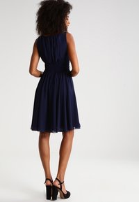 Robe bleu marine avec une jupe évasée, une taille froncée et sans manches. Accompagnée de sandales à talons noirs. Texture lisse.