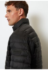 Chaqueta de plumas negra con cuello alto y textura acolchada. Presenta un parche con logo en el lado izquierdo y un diseño elegante y ligero.