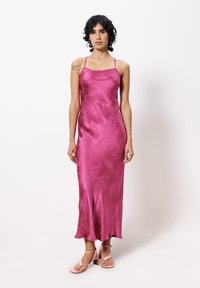 Zijdeachtige roze slipdress met dunne bandjes, met een subtiel bloemenpatroon; vloerlange met een lichte flare aan de zoom. Gecombineerd met roze hoge hakken.