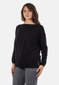 Donna che indossa un maglione nero a maniche lunghe e jeans grigi, in piedi con una mano sulla vita contro uno sfondo bianco semplice.