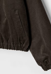 Giacca marrone in velluto a coste con trama a costine, polsini elastici e dettaglio tasca laterale. Il tessuto presenta righe verticali.