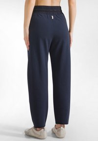 Osoba w luźnych granatowych spodniach i jasnobeżowych trampkach stoi tyłem na jednolitym białym tle.