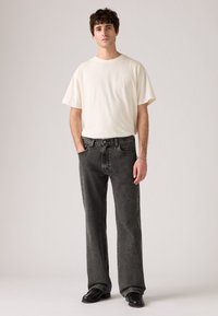 Jonge man die met zijn handen in zijn zakken staat, gekleed in een eenvoudig wit T-shirt, vervaagde zwarte jeans, zwarte schoenen en een zilveren armband.