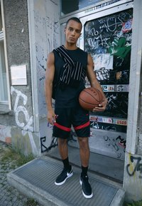 Sort ærmeløs skjorte med hvide striber, sorte shorts med røde detaljer og sorte sneakers. Holder en basketball og står op ad en væg dækket af graffiti.