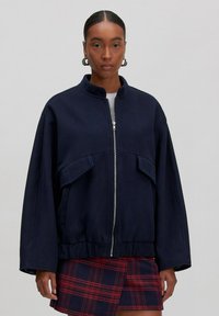 Marineblaue, übergroße Zip-Jacke mit hohem Kragen, großen Fronttaschen und elastischem Saum, kombiniert mit einem karierten Rock. Weiche Textur.