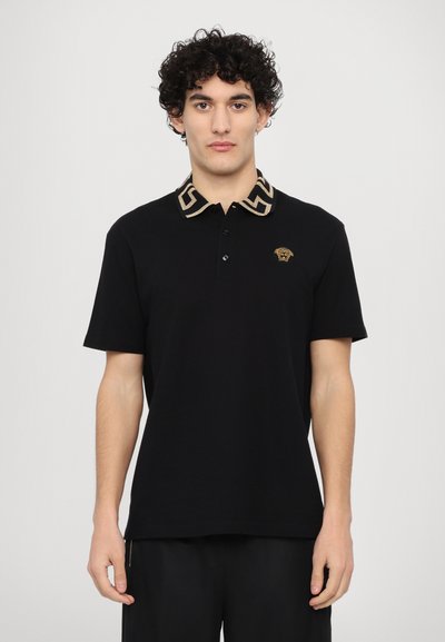 Camisa polo negra de manga corta, con un collar a rayas en oro y negro y un logotipo dorado en el pecho. Material de algodón.