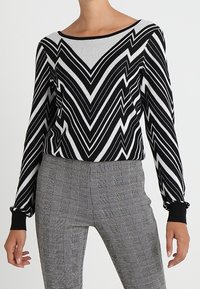 Schwarzer und weißer Zickzack-musterter Pullover mit langen Ärmeln, rundem Ausschnitt und gerippten Bündchen, kombiniert mit grauen karrierten, hochtaillierten Hosen.
