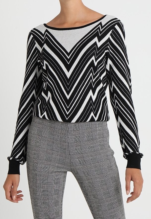 Schwarzer und weißer Zickzack-musterter Pullover mit langen Ärmeln, rundem Ausschnitt und gerippten Bündchen, kombiniert mit grauen karrierten, hochtaillierten Hosen.