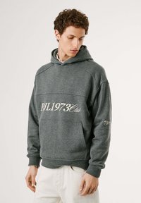 Felpa grigia con cappuccio e tasca anteriore, con il testo bianco "PJL1973Club" e un piccolo logo sulla manica. Tessuto morbido e polsini a costine.