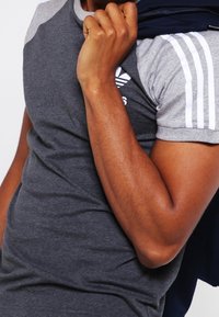 Hombre con una camiseta gris de Adidas sosteniendo una chaqueta azul marina sobre su hombro frente a un fondo liso.