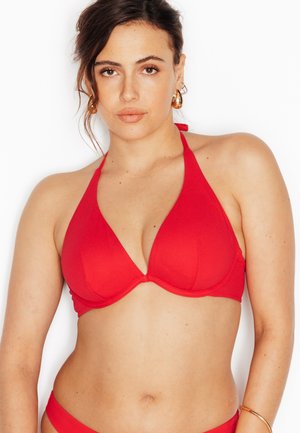 Donna con capelli castani che indossa orecchini a cerchio dorati e un top bikini a fascia rosso su sfondo bianco uniforme.