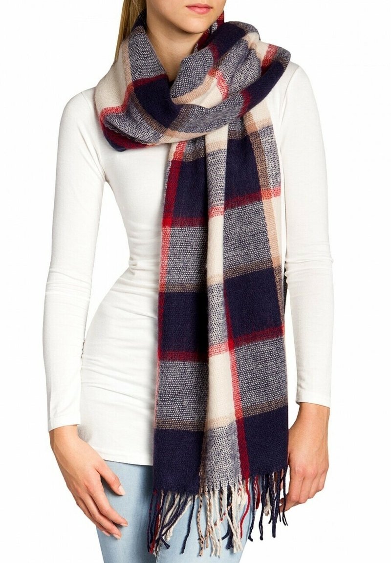 Femme portant une grande écharpe épaisse à carreaux aux couleurs bleu marine, rouge, beige et gris sur un haut blanc à manches longues et un jean bleu clair.