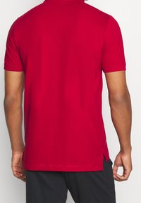 Polo rouge en tissu lisse, doté d'un col classique et de manches courtes, avec des fentes latérales à l'ourlet pour faciliter les mouvements.