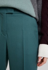 Pantalons ajustés teal avec une texture lisse et des plis à l'avant. Un pull en tricot gris, bordeaux et blanc est partiellement visible en haut.