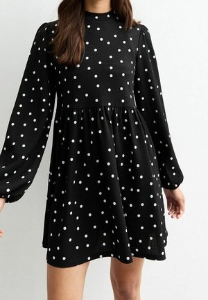 Kobieta w czarnej sukience w białe groszki, z długimi bufkami i talią zbieraną, stojąca na jasnym, jednolitym tle.