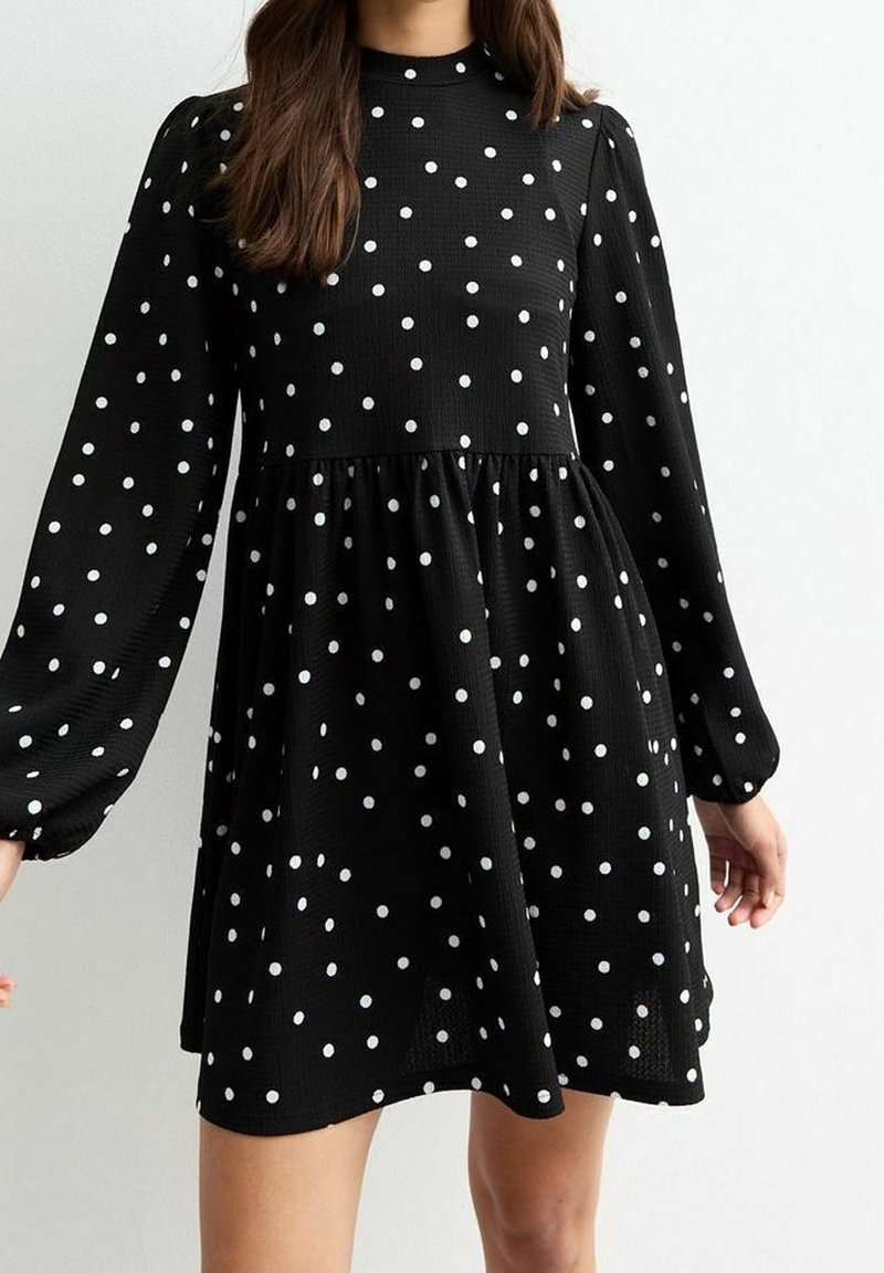 Femme portant une robe noire à pois blancs, manches longues bouffantes et taille froncée, debout devant un fond clair uni.