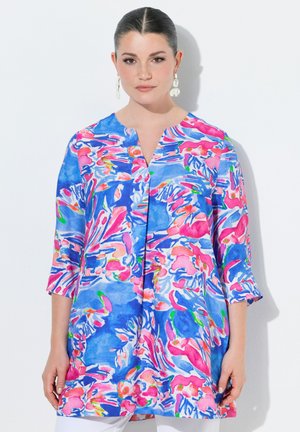 Frau trägt eine lebhafte, locker sitzende Tunika mit abstrakten Blumenmustern in Blau, Pink und Weiß sowie dreiviertellangen Ärmeln.