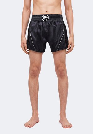 Bărbat purtând pantaloni scurți sport negri cu un logo de șarpe pe talie, stând desculț pe un fundal simplu.