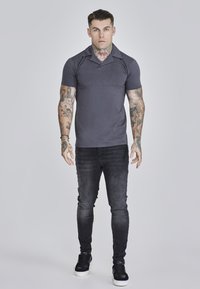 Camisa polo gris con cuello, mangas cortas y un diseño ajustado, combinada con jeans skinny oscuros y zapatillas negras.