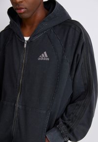 Black hooded zip-up sweatshirt with Adidas logo, featuring ribbed cuffs and three stripes design on sleeves. Textured fabric, relaxed fit.