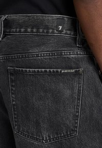 Jeans in denim nero con una tasca posteriore dotata di un piccolo etichetta. Il tessuto presenta una texture sbiadita con cuciture a contrasto lungo le cuciture.